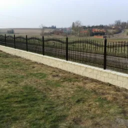 Eleganckie, kute ogrodzenie w kolorze czarnym, osadzone na niskim murku z jasnych bloczków, na tle wiejskiego krajobrazu. Widok z perspektywy trawnika.