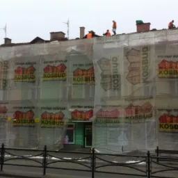 Budynek w trakcie ocieplania, osłonięty siatką z logo firmy KOSBUD, pracownicy na dachu. Widoczne są sąsiednie budynki i ulica.