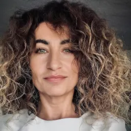 Portret kobiety z burzą kręconych, brązowo-blond włosów, zielone oczy, neutralny wyraz twarzy, na ciemnoszarym tle.