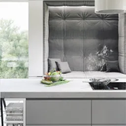 Nowoczesna kuchnia z szarą tapicerowaną wnęką do siedzenia, owocami w szklanej misce, patelnią na płycie indukcyjnej i półką z książkami.