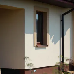 Fragment elewacji domu z oknami w brązowych ramach, rynną i kolumną. Jasna elewacja z ciemnobrązowym cokołem. Widoczne detale wykończenia.