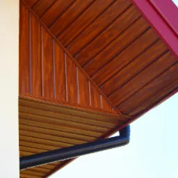 Drewniana podbitka dachowa z czerwonymi elementami wykończeniowymi i czarną rynną, widoczna z dołu, przylegająca do białej ściany budynku.