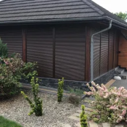 Brązowe rolety zewnętrzne na altanie z ciemnym dachem, otoczonej kamieniami i roślinnością, w tle widoczny budynek z zielonym żywopłotem.