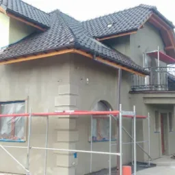 Dom jednorodzinny w trakcie tynkowania elewacji, widoczne rusztowania, okna zabezpieczone folią, fragmenty elewacji z dekoracyjnymi elementami architektonicznymi.