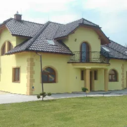 Żółty dom jednorodzinny z ciemnobrązową dachówką, widok z trawnika, z elementami dekoracyjnymi elewacji imitującymi kamień.