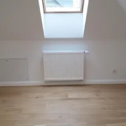 Poddasze po remoncie z oknem dachowym, białymi ścianami, grzejnikiem i drewnianą podłogą z jasnych paneli.