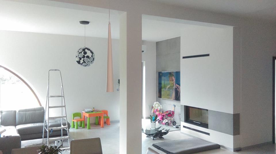 Wnętrze nowoczesnego salonu po remoncie z białą ścianą, kominkiem obłożonym szarym betonem, schodami drabinkowymi i designerskimi lampami wiszącymi.
