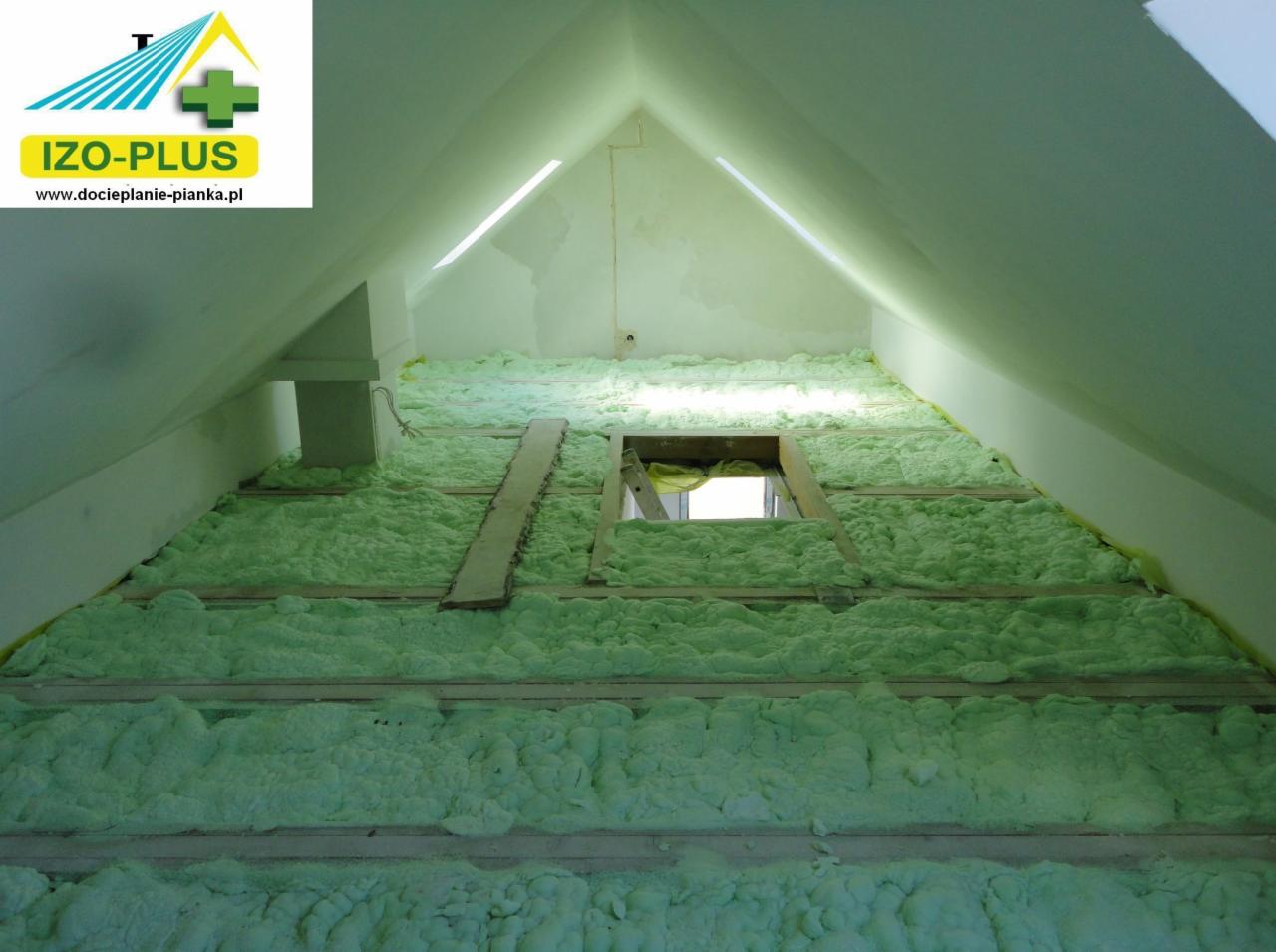Poddasze z widoczną zieloną pianką izolacyjną pomiędzy drewnianymi belkami, oświetlone światłem dziennym wpadającym przez okno dachowe.