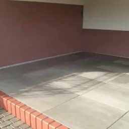 Wykończony taras z szarych płyt betonowych, obrzeże z cegły klinkierowej. Widoczne cienie rzucane przez konstrukcję nad tarasem. Ściana w kolorze bordowym.