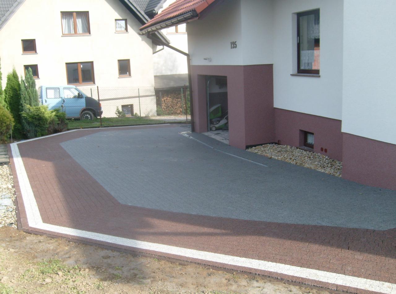 Ułożona kostka brukowa w kolorach szaro-brązowym tworząca podjazd do domu z numerem 125. Krawędzie wykończone białym pasem. W tle budynki i drzewa.