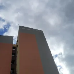 Wysoki budynek z pomarańczowymi i szarymi panelami, widziany z dołu na tle nieba z chmurami. Widoczny fragment dachu i okna.