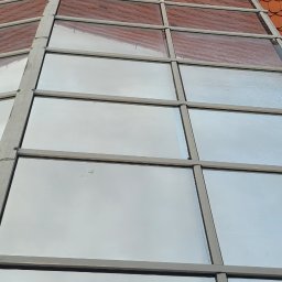 NO LIMIT Damian Krzesak - Szklany dach z metalową konstrukcją i pomarańczową dachówką w tle. Widoczne ślady po umyciu, sugerujące niedawne czyszczenie. Ujęcie z dołu, podkreślające geometrię.