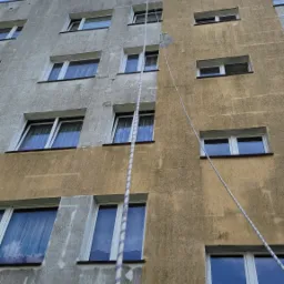 Budynek z elewacją w połowie wyczyszczoną, widoczna różnica między brudną a czystą częścią. Lina pionowo wzdłuż ściany. Ujęcie z dołu, niebo w tle.