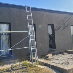 NO LIMIT Damian Krzesak - Świeżo pomalowana, grafitowa elewacja budynku z oknami zabezpieczonymi folią. Widoczne rusztowania i drabina, sugerujące trwające prace wykończeniowe.
