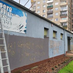 NO LIMIT Damian Krzesak - Szara ściana budynku z częściowo zamalowanym graffiti, drabina oparta o ścianę. Widoczny fragment reklamy i okna z kratami. W tle blok mieszkalny.