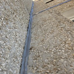 NO LIMIT Damian Krzesak - Szczelina dylatacyjna wypełniona szarą masą uszczelniającą na elewacji budynku z fakturą kamyczkową, widok z dołu, fragment liny zabezpieczającej w górnej części kadru.