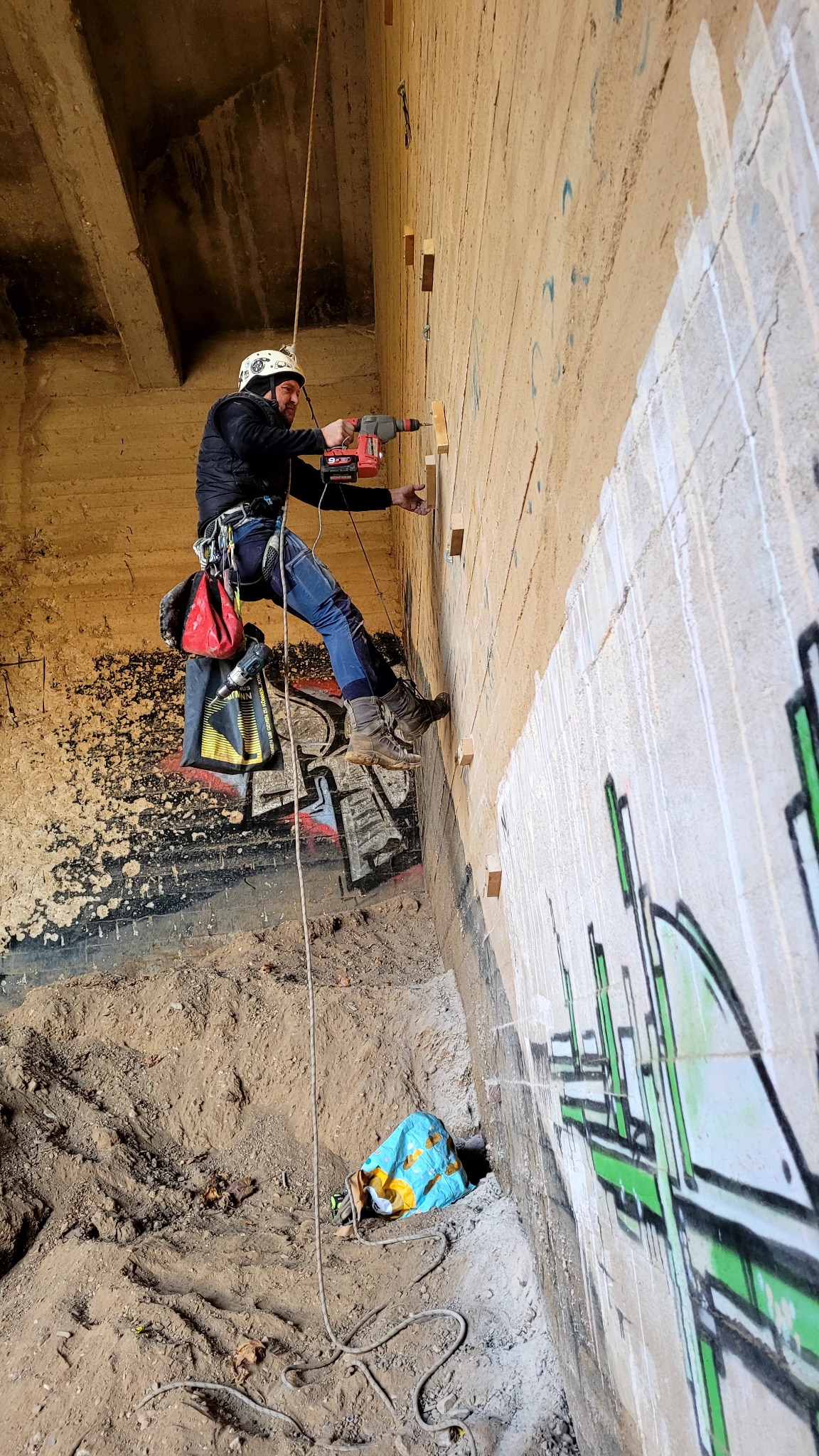 Pracownik na linie wierci otwory w betonowej ścianie z graffiti, zabezpieczony kaskiem i uprzężą. Widoczne drewniane elementy mocowane do ściany.