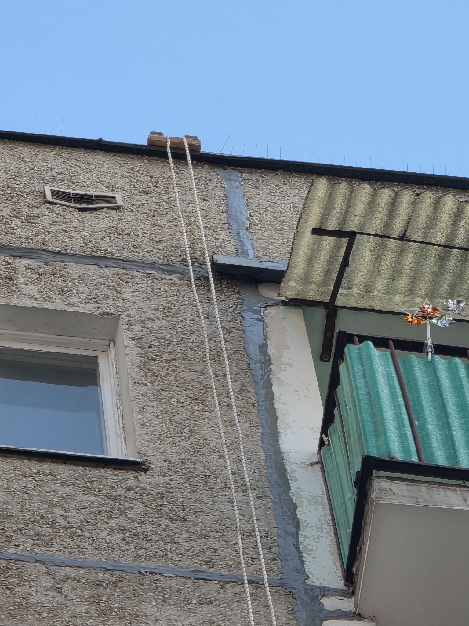 Szara elewacja budynku z balkonem, zabezpieczona kolcami przeciw ptakom. Widoczna lina przymocowana do dachu, sugerująca prace na wysokości.