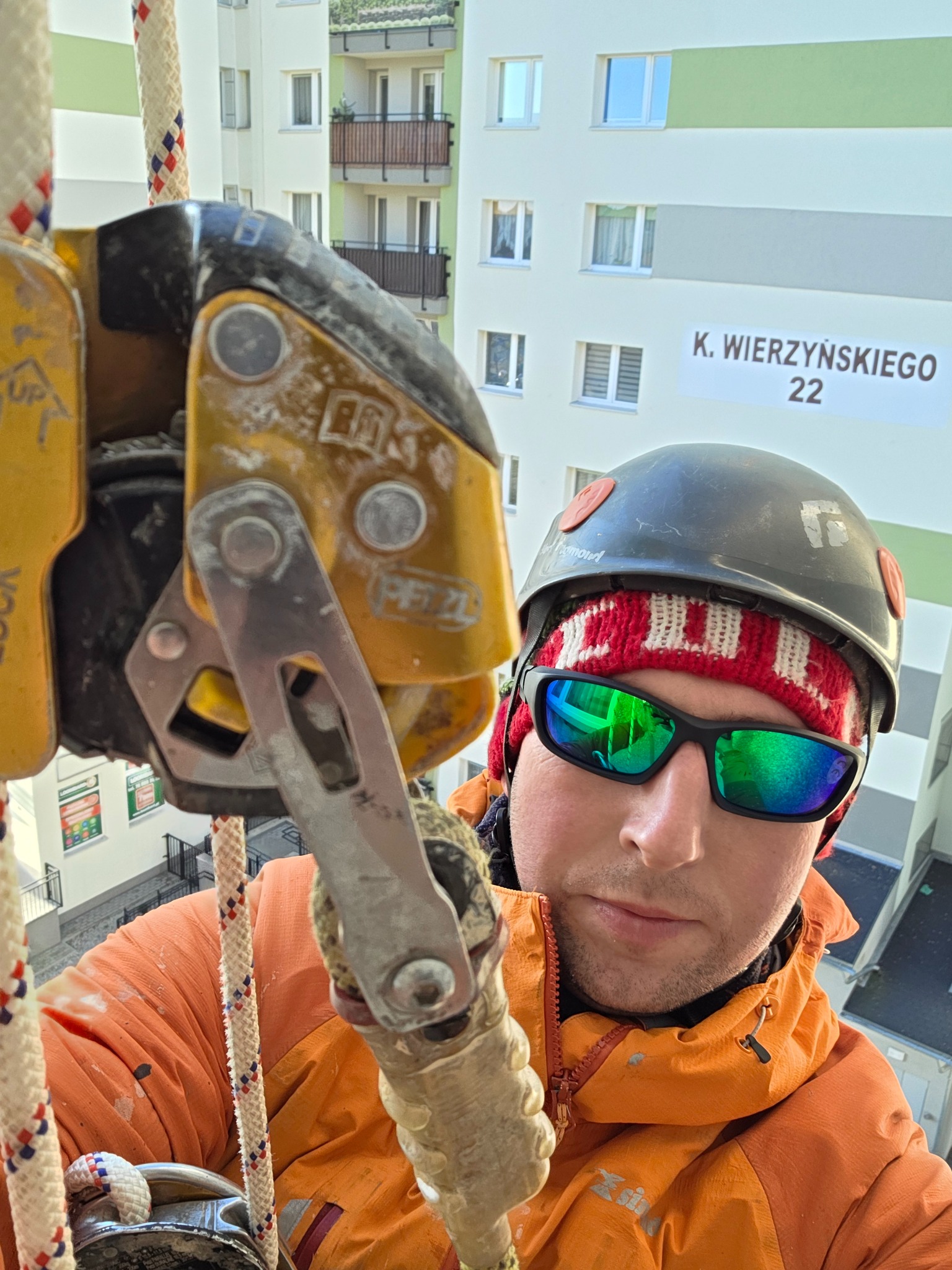 Pracownik w kasku i okularach, zabezpieczony linami, w trakcie pracy na wysokości przy budynku z adresem K. Wierzyńskiego 22. Widok z dołu, sprzęt alpinistyczny.