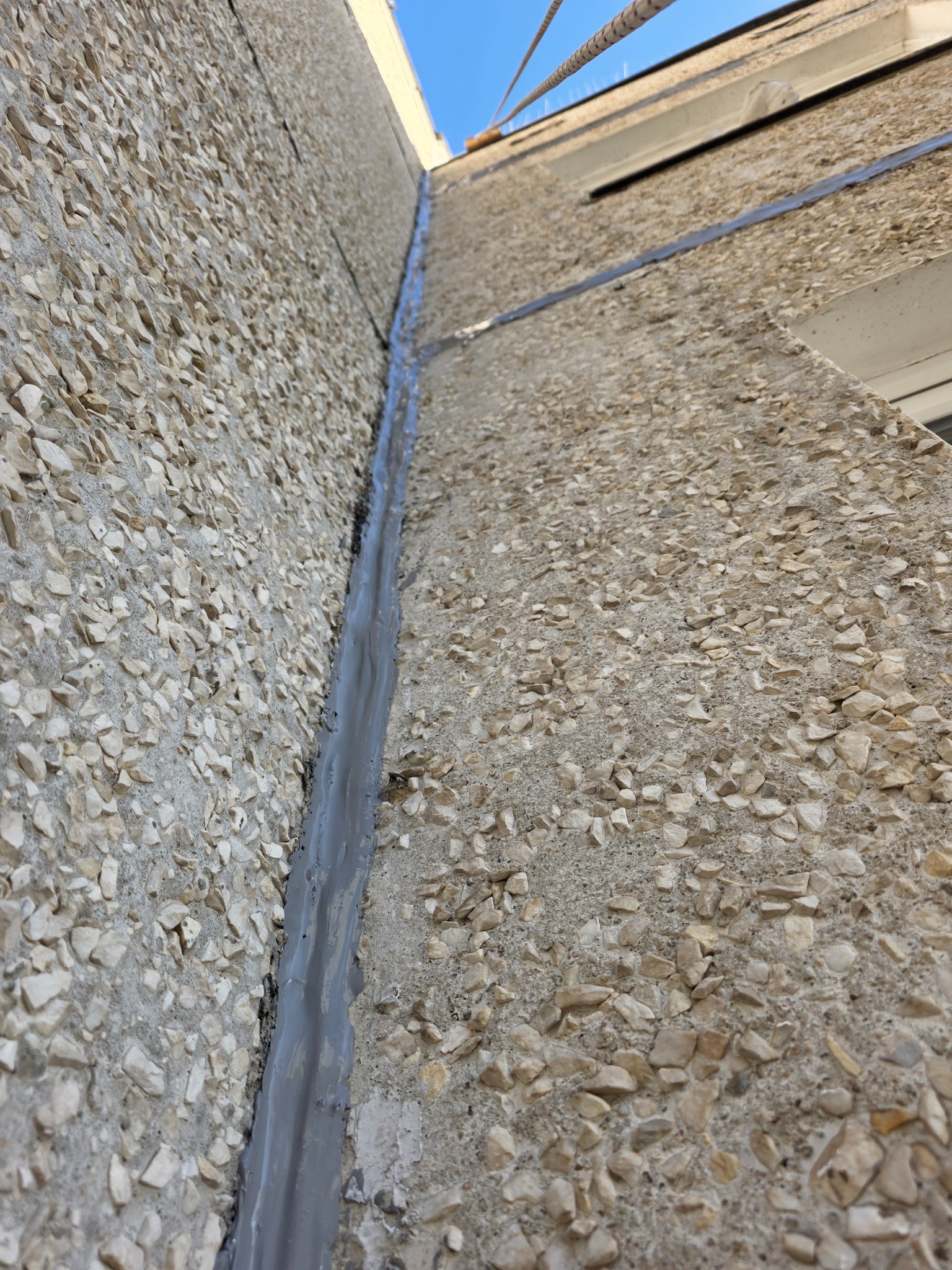 Szczelina dylatacyjna wypełniona szarą masą uszczelniającą na elewacji budynku z fakturą kamyczkową, widok z dołu, fragment liny zabezpieczającej w górnej części kadru.