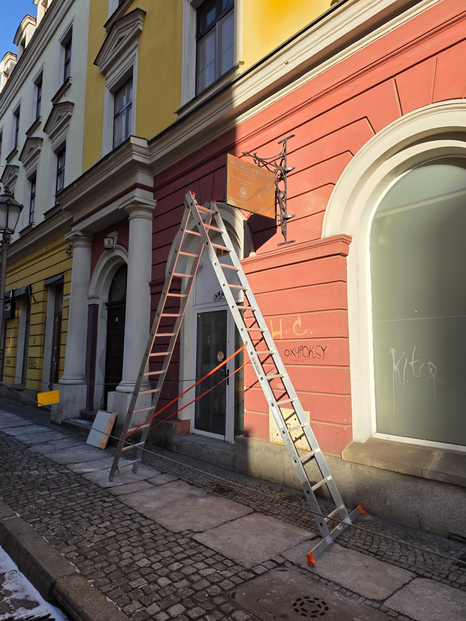 Rozstawiona drabina oparta o czerwoną elewację budynku z widocznymi napisami i szyldem. Kamienna kostka brukowa na chodniku. Prace na wysokości.
