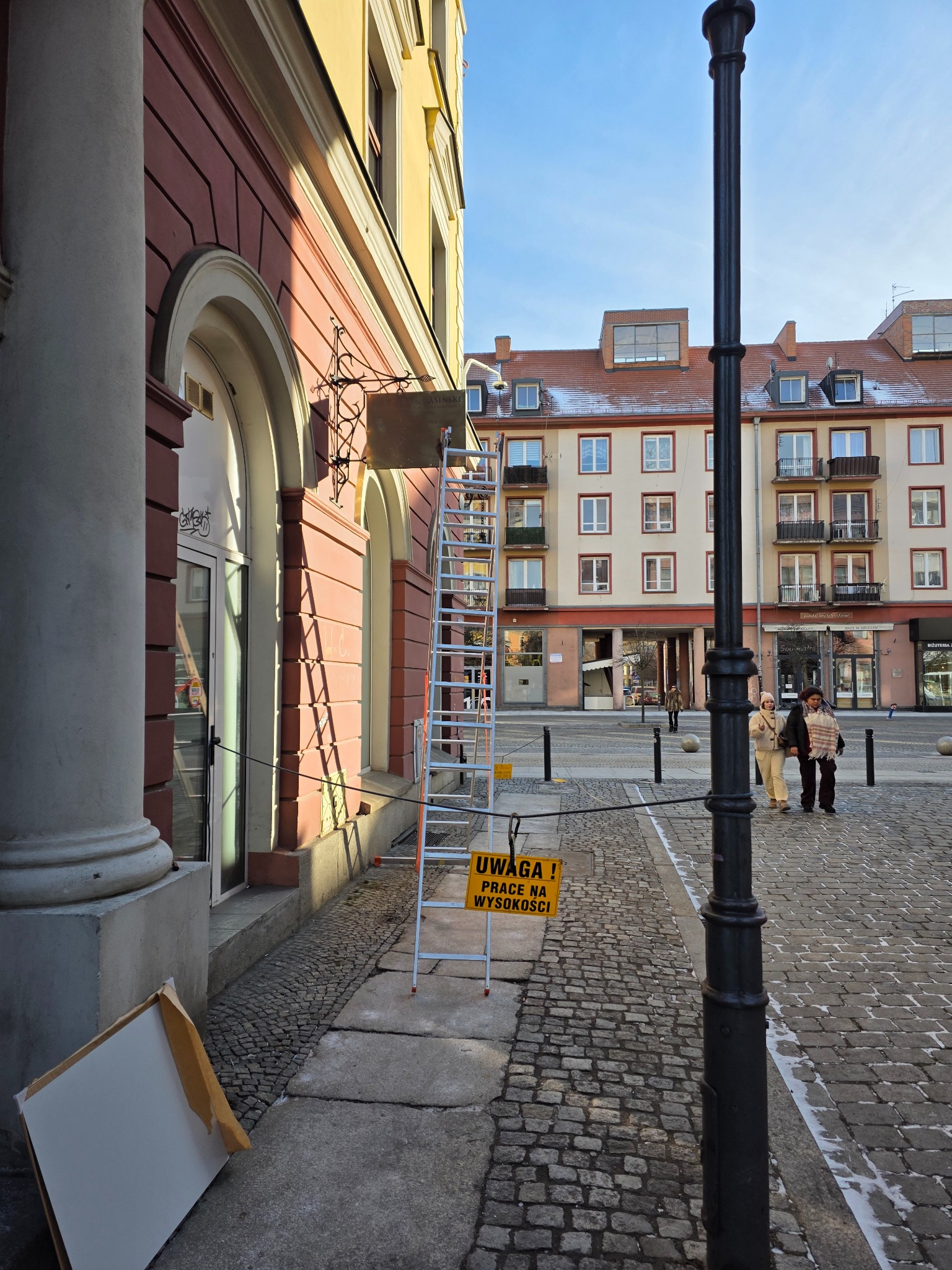 Aluminiowa drabina oparta o budynek z szyldem, tabliczka ostrzegawcza 'Prace na wysokości', kolumna i fragment elewacji w słoneczny dzień na brukowanym placu.