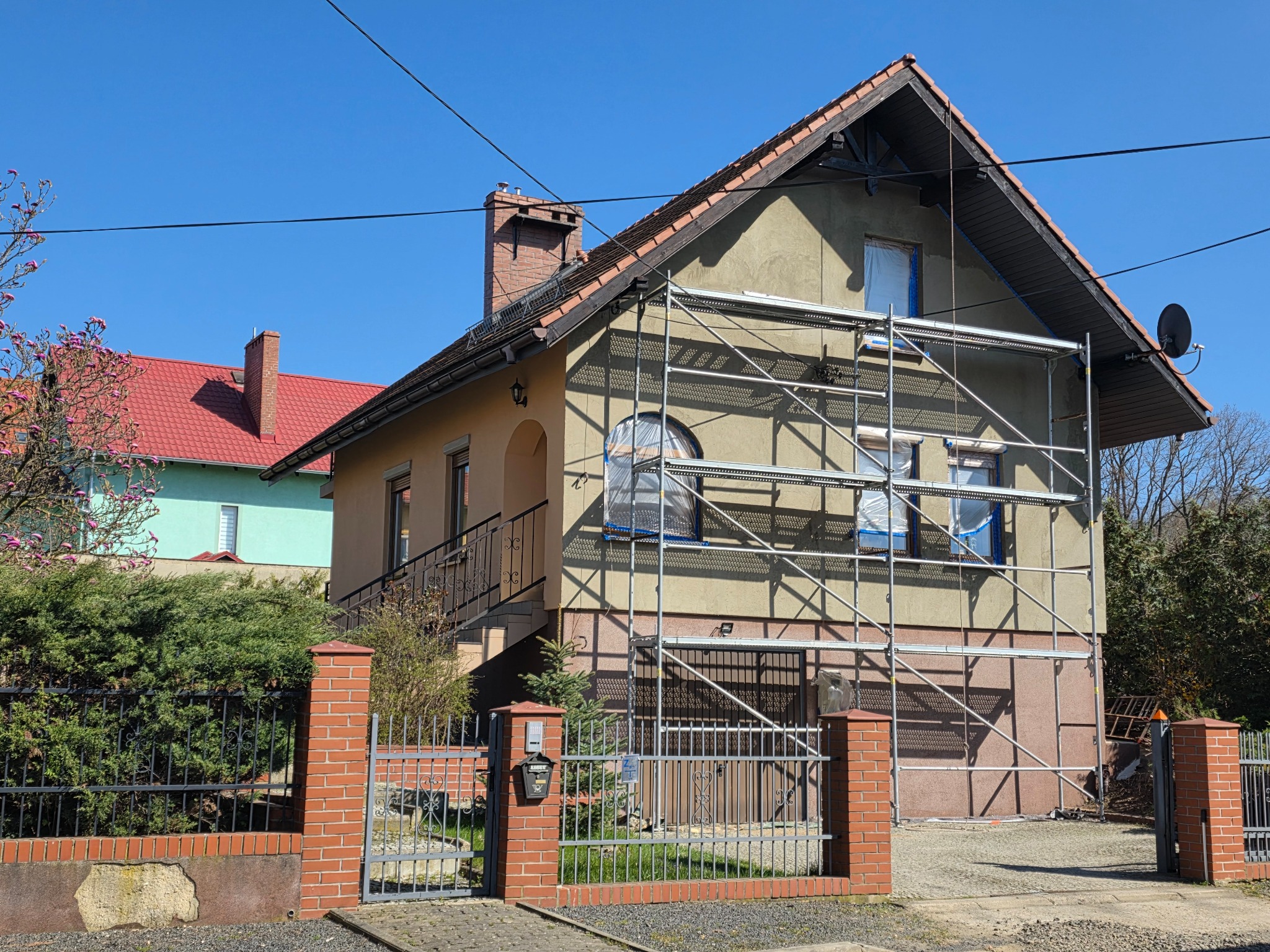 Dom jednorodzinny w trakcie prac elewacyjnych, z rusztowaniem i zabezpieczonymi oknami. Widoczny fragment ogrodzenia z cegły i metalu. Słoneczny dzień.