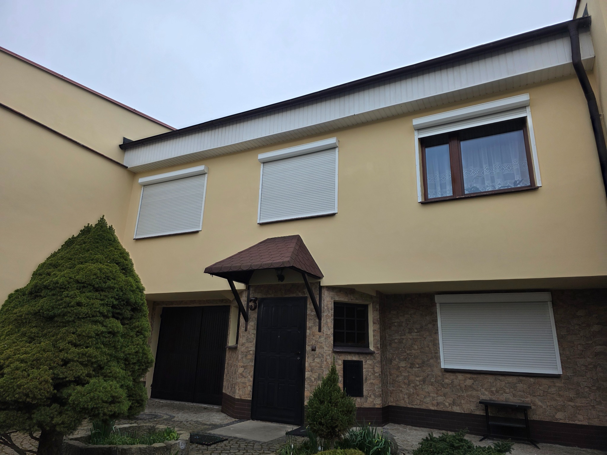Dwukondygnacyjny dom z beżową elewacją, brązowym dachem i szarymi roletami zewnętrznymi. Część fasady wykończona kamieniem. Widoczne krzewy i drzewa.