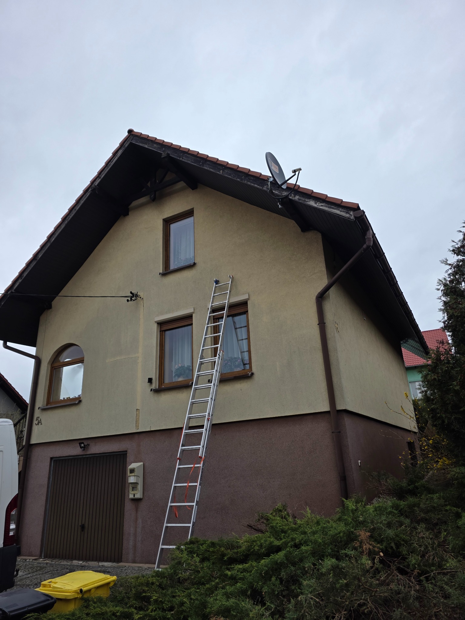 Dwukondygnacyjny dom z beżowo-brązową elewacją. Aluminiowa drabina oparta o ścianę, widoczne okna z brązowymi ramami, brązowe rynny i antena satelitarna na dachu.