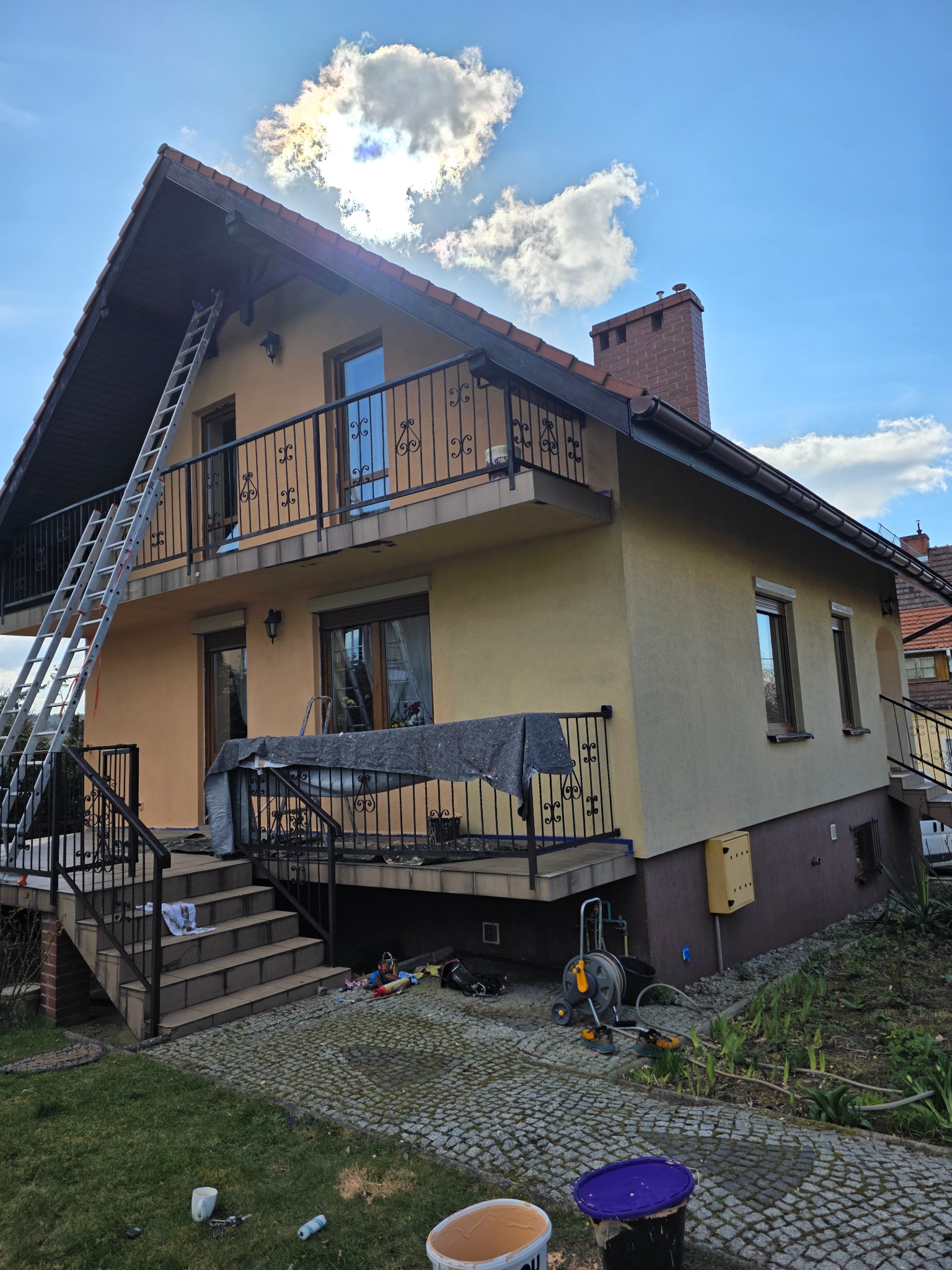 Dom z balkonem w trakcie malowania elewacji. Rozstawiona drabina, widoczne narzędzia i farby. Część elewacji pomalowana na ciemniejszy odcień.