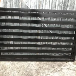 Czarna, metalowa brama z poziomymi szczeblami, zawieszona na zawiasach, na tle blaszanej ściany i betonowej posadzki.