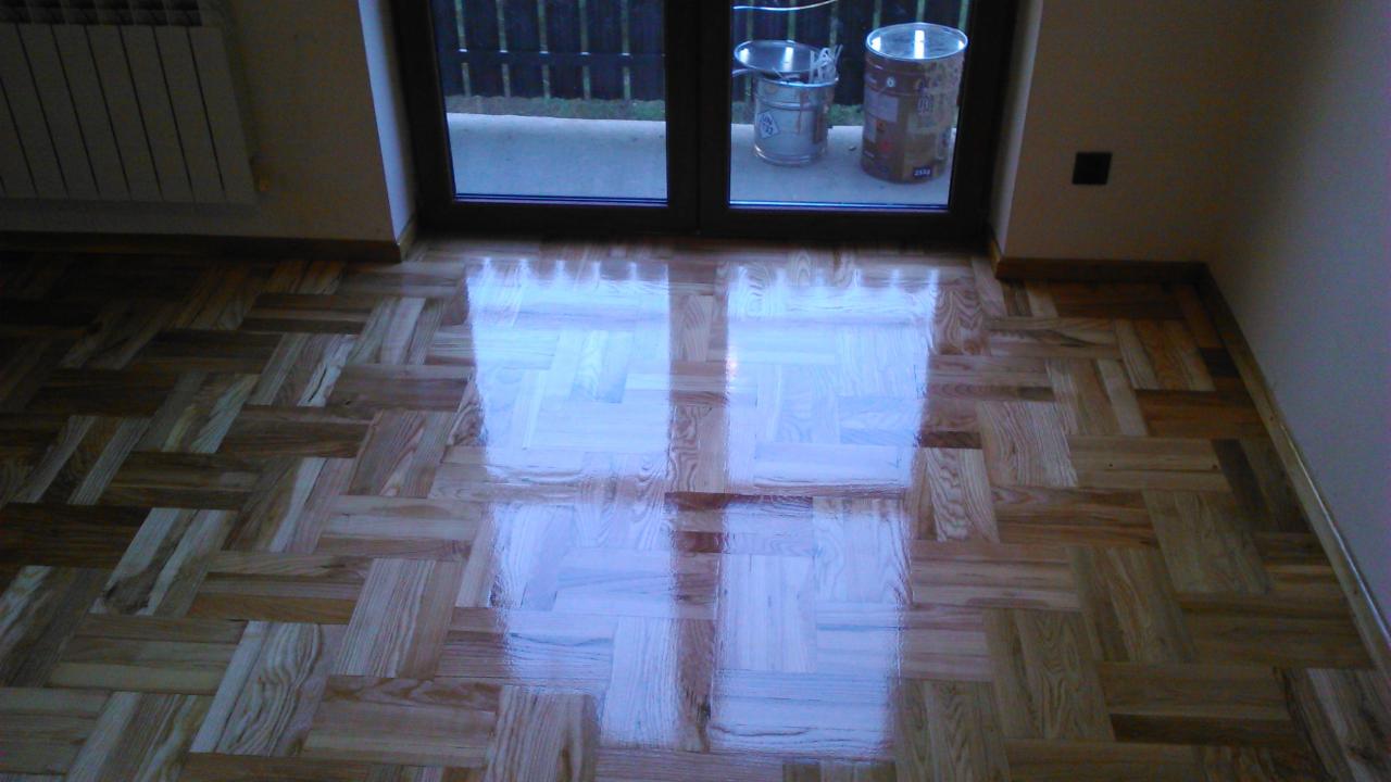 Parkiet ułożony w jodełkę po renowacji, z widocznym połyskiem świeżo nałożonego lakieru, odbijający światło z okna balkonowego; widoczne puszki po lakierze.