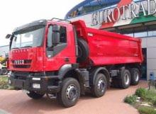 Czerwona wywrotka Iveco Eurotrakker na tle budynku z logo Eurotrade, zaparkowana na kostce brukowej. Widok z boku, lekko od dołu. Opona zapasowa za kabiną.