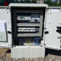 Otwarta szafa elektryczna z widocznymi bezpiecznikami, przełącznikami i okablowaniem, zamontowana na zewnątrz budynku, na tle zielonego ogrodzenia.