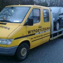 Żółta laweta z napisem 'Pomoc Drogowa' przewozi dwa samochody osobowe na tle drzew. Laweta ma dodatkowe oświetlenie i widoczne numery telefonów.