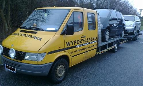 Żółta laweta z napisem 'Pomoc Drogowa' przewozi dwa samochody osobowe na tle drzew. Laweta ma dodatkowe oświetlenie i widoczne numery telefonów.