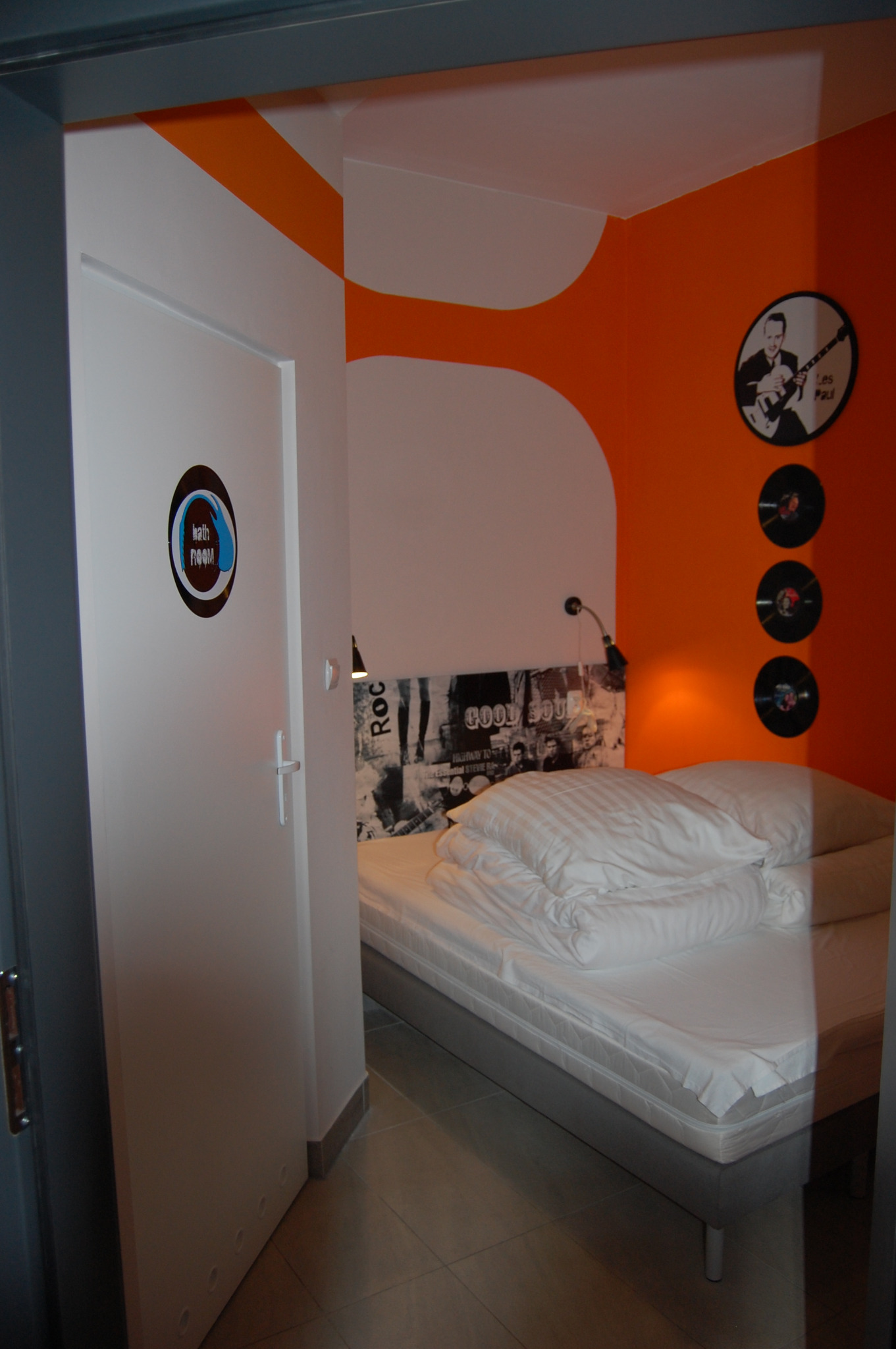 Nowoczesny pokój hotelowy z łóżkiem, pomarańczowo-białymi ścianami z motywem muzycznym, widok zza uchylonych drzwi z oznaczeniem 'bath room'.