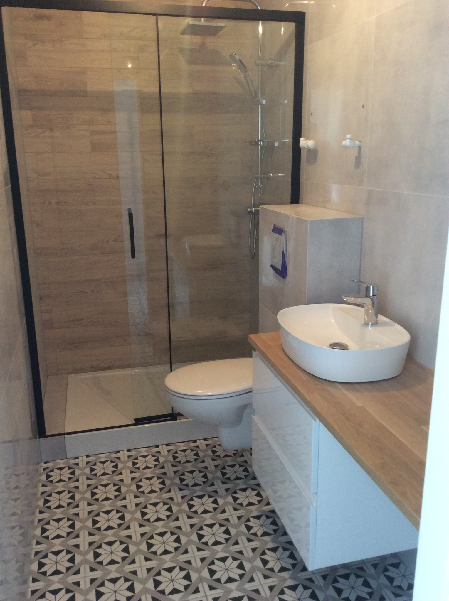 Nowoczesna łazienka z kabiną prysznicową w czarnej ramie, wiszącą umywalką na drewnianym blacie i wzorzystą podłogą.