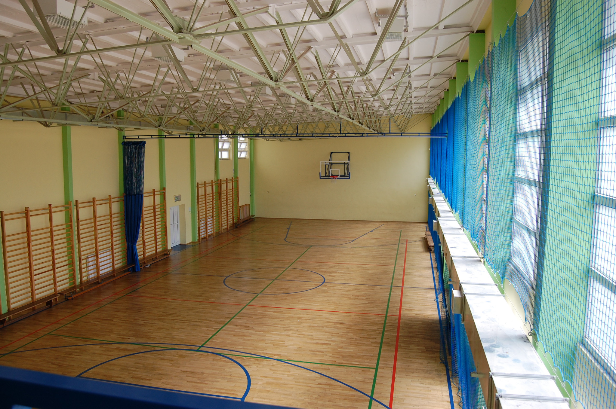 Wnętrze przestronnej sali gimnastycznej z drewnianą podłogą z namalowanymi liniami boisk, metalową konstrukcją dachu i siatką okienną w kolorze niebieskim.