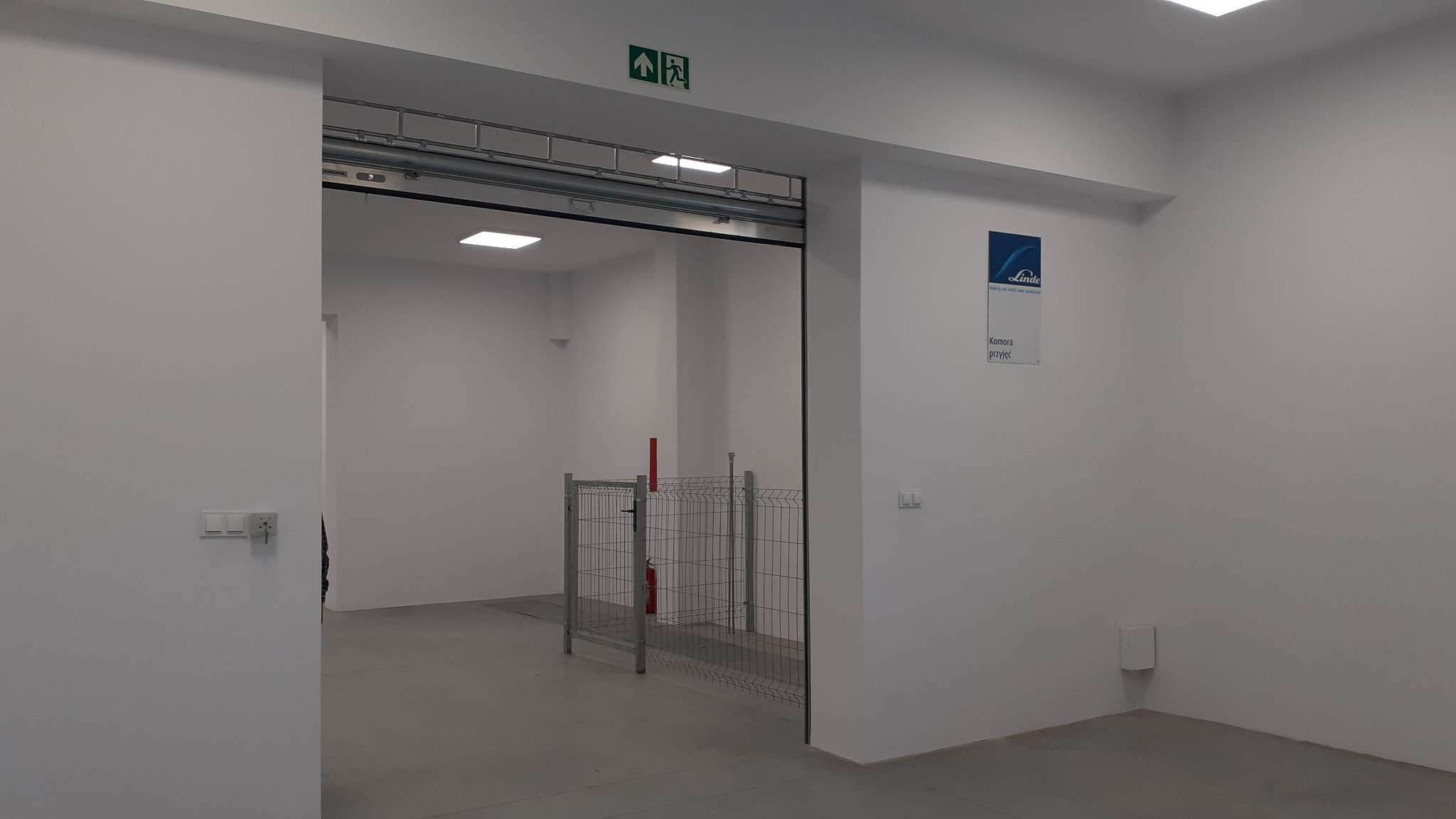 Wnętrze hali magazynowej z bramą przesuwną, widoczne zabezpieczenia w postaci metalowej klatki, znak ewakuacyjny i logo firmy Linde na ścianie.