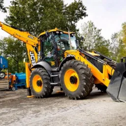 Żółto-czarna koparko-ładowarka JCB na betonowym placu, z zielonymi drzewami w tle i niebieskimi maszynami rolniczymi po lewej stronie.