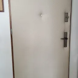 Drzwi wejściowe obite kremową, pikowaną tkaniną z drewnianą ramą i srebrnymi okuciami, widoczne zawiasy i wizjer.