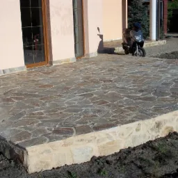 Wykończony taras z nieregularnych płyt kamiennych na podwyższonej wylewce, widoczny skuter i osoba w tle.