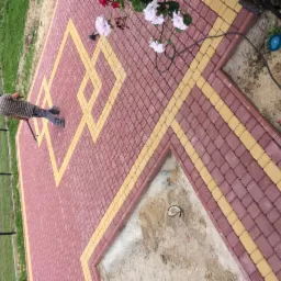 Pracownik ubija czerwoną kostkę brukową z żółtymi akcentami, tworząc geometryczny wzór na ścieżce w ogrodzie, widoczne niedokończone fragmenty nawierzchni.