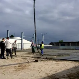 Betonowanie terenu pod budowę hali. Robotnicy w trakcie wylewania betonu z użyciem pompy. Widoczne stalowe elementy konstrukcyjne i szalunki.