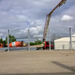 Budowa hali: betonowanie fundamentów. Widoczny betonowóz z pompą, stalowe słupy konstrukcyjne i pracownik wyznaczający punkty. Pochmurne niebo.