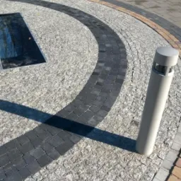 Okrągła nawierzchnia z jasnoszarej kostki granitowej, obramowana ciemniejszą kostką. Widoczne słupki oświetleniowe i fragment szarej kostki brukowej w tle.