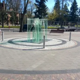 Plac miejski z kostki brukowej z okrągłą, ozdobną instalacją z zielonego szkła i metalu. Ławki i drzewa w tle. Słoneczny dzień.