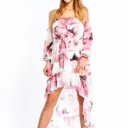 Blondwłosa kobieta w zwiewnej, kwiecistej sukience z asymetrycznym dołem i czarnych sandałach na obcasie z futerkiem, pozująca na białym tle.