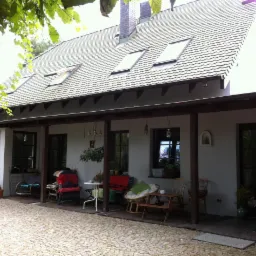 Dom jednorodzinny z szarą dachówką, oknami dachowymi i zadaszonym tarasem z meblami ogrodowymi, widok z zewnątrz na kamienny podjazd.